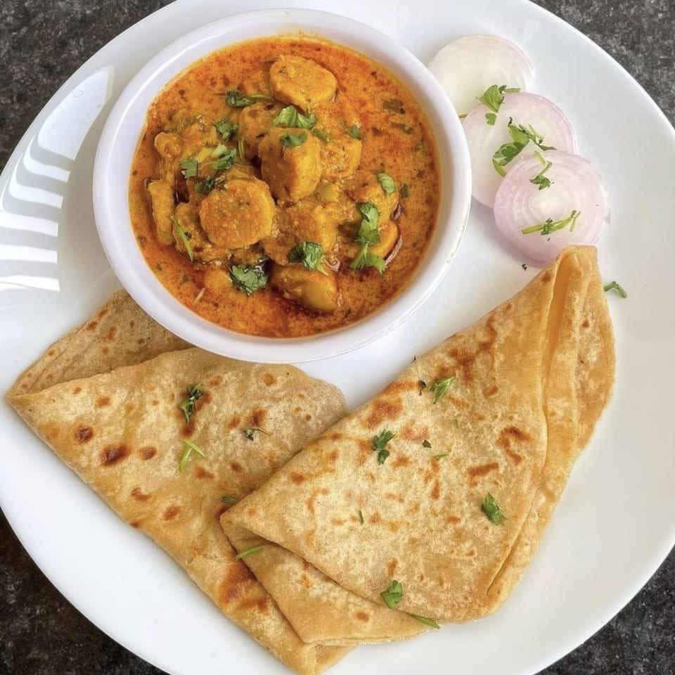 A white plate with a bowl of chicken curry, sliced red onions, and two folded flatbreads.