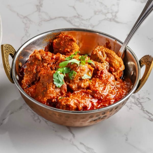 A metal bowl filled with a rich, red curry, garnished with fresh green cilantro, on a marble surface.