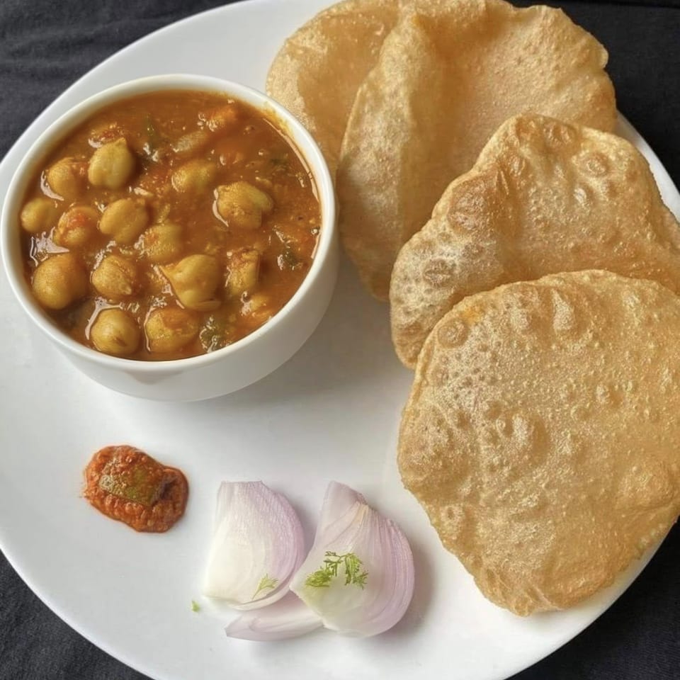 A white plate holds chole bhature, with a bowl of chickpea curry, fried bread, onions, and chutney.