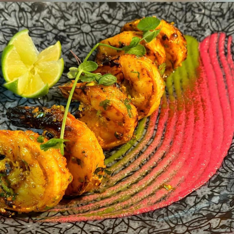 Grilled shrimp with lime wedges, microgreens, and vibrant pink and green sauces on a patterned plate.